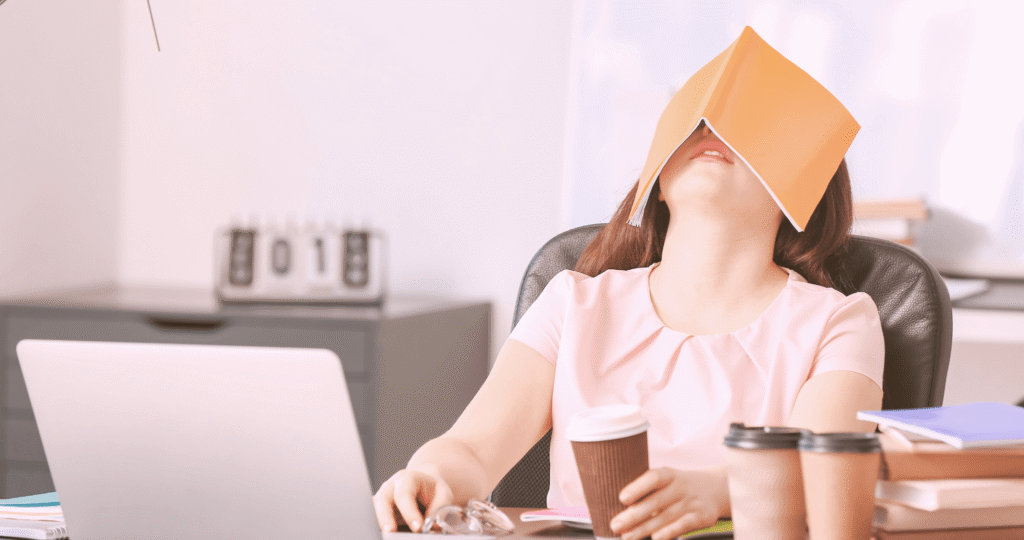 Mulher sentada em uma cadeira de escritório com um caderno cobrindo o rosto, cercada por copos de café em uma mesa, sugerindo exaustão causada por trabalho excessivo, representando a neurosciência da comunicação quando o cérebro entra em alerta.